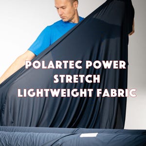 Pode incluir: Uma pessoa segurando um grande pedaço de tecido leve Polartec Power Stretch azul marinho. O tecido é drapeado para mostrar sua textura e caimento. O texto "POLARTEC POWER STRETCH LIGHTWEIGHT FABRIC" é sobreposto à imagem em branco.