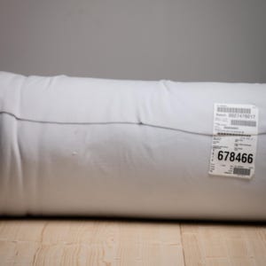 Op de afbeelding: Een wit, cilindrisch kussen op een licht houten oppervlak. Een label met het nummer "678466" is bevestigd. De achtergrond is effen grijs. Het kussen lijkt van een zacht materiaal te zijn.