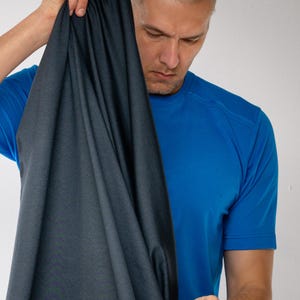 Pode incluir: Uma pessoa segurando um tecido cinza escuro, possivelmente roupa esportiva. O tecido está drapeado e parece macio. A pessoa está vestindo uma camisa esportiva azul. O fundo é uma parede branca lisa.