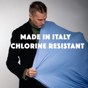 Può includere: Una persona tiene in mano un tessuto azzurro. Il tessuto è teso, rivelando la sua consistenza. Il testo "MADE IN ITALY CHLORINE RESISTANT" è stampato sul tessuto con lettere bianche. La persona indossa una giacca nera e un dolcevita verde.
