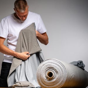 May include: A person inspecting a piece of grey fabric. A large roll of grey fabric is on a wooden surface. The fabric is a neutral colour, and the image is well-lit, highlighting the texture and quality of the material. The fabric appears to be a knit or jersey material.
