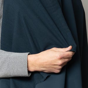 Könnte beinhalten: Nahaufnahme einer Hand, die ein großes Stück dunkelblaugrünen Stoff hält. Der Stoff wirkt weich und drapiert sich in Falten. Die Person trägt ein graues Langarmshirt.