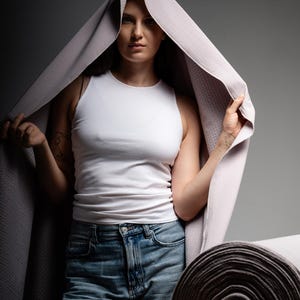 Peut inclure: Une personne en débardeur blanc et jean bleu tient une couverture violet clair au-dessus de sa tête. Une section enroulée de la couverture est visible à droite. La couverture a un motif texturé.