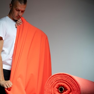 May include: A person holding up a vibrant orange fabric roll. The fabric is a solid, bright color, and appears to be a medium-weight material. A second roll of the same fabric is visible on a wooden surface.
