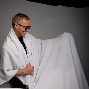 Op de afbeelding: Een lichtgrijze stof gedrapeerd over een persoon met een zonnebril en een zwart shirt. De stof wordt omhoog gehouden, waardoor plooien en schaduwen ontstaan. De achtergrond is neutraal grijs.