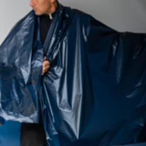 May include: A person is holding up a large roll of navy blue fabric. The fabric is shiny and appears to be waterproof. A roll of the same fabric is on a wooden surface in the foreground.