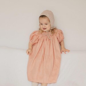 Puede incluir: Un vestido color melocotón con mangas abullonadas y detalles de nido de abeja en el escote, combinado con un gorro texturizado a juego que se ata bajo la barbilla y calcetines claros. La prenda presenta una silueta holgada y fluida.