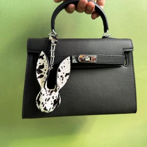 Handmade bunny leather bag charm made from horsehair and lambskin, shown hanging on a black handbag. Luxury purse charm and statement handbag charms featuring hair-on leather texture, designed as a unique bag accessory.