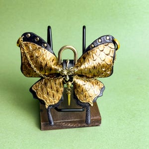 May include: A decorative butterfly-shaped object with gold and black accents. The wings have a textured, scale-like pattern. A gold chain and a gold-colored lock are attached to the butterfly. The object is displayed on a small wooden stand.