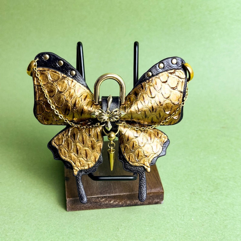 May include: A decorative butterfly-shaped object with gold and black accents. The wings have a textured, scale-like pattern. A gold chain and a gold-colored lock are attached to the butterfly. The object is displayed on a small wooden stand.