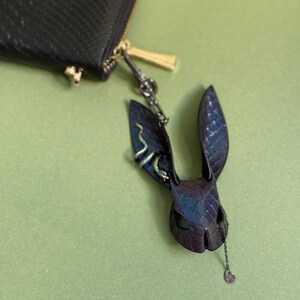 May include: A black leather wallet with a gold zip is attached to a dark blue and purple rabbit head keyring. The keyring has a snake design on one ear and a dangling chain with a purple gem.