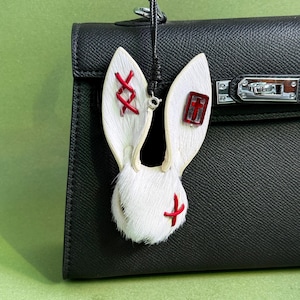 May include: A white rabbit ear-shaped bag charm with red stitching and accents. The charm is made of fur and leather, with a red cross and a red cross-shaped design. The charm is hanging from a black leather bag.
