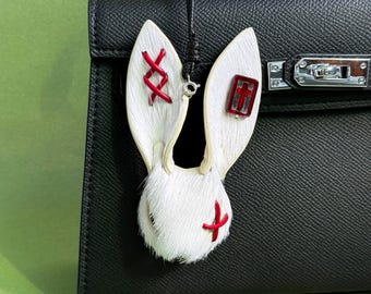 May include: A white rabbit ear-shaped bag charm with red stitching and accents. The charm is made of fur and leather, with a red cross and a red cross-shaped design. The charm is hanging from a black leather bag.