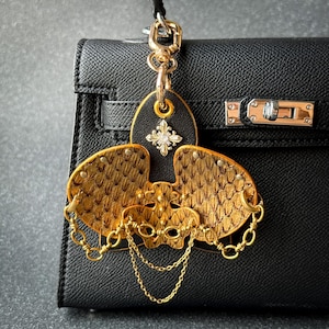 May include: A black leather handbag featuring a gold-toned metal bag charm shaped like an orchid. The charm has textured gold petals, a central silver-toned fleur-de-lis emblem, and delicate gold chains draped across the lower section.