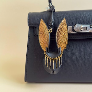 Peut inclure: Un sac à main en cuir noir avec un charm en forme de chauve-souris. Le charm présente des ailes texturées dorées et marron, un corps noir et des chaînes dorées pendantes. Le sac a un fermoir argenté et une poignée noire.