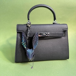 Blue-purple rainbow leather bunny bag charm hanging on a black handbag. Handmade lambskin and snakeskin-textured rabbit head charm with iridescent blue-purple color shifts. Long ears, carved contours, and a small dangling pendant chain showcase detailed craftsmanship. Works as a leather bag charm, purse charm, keychain charm, or car mirror charm. Photographed against a green background to highlight the rainbow sheen.