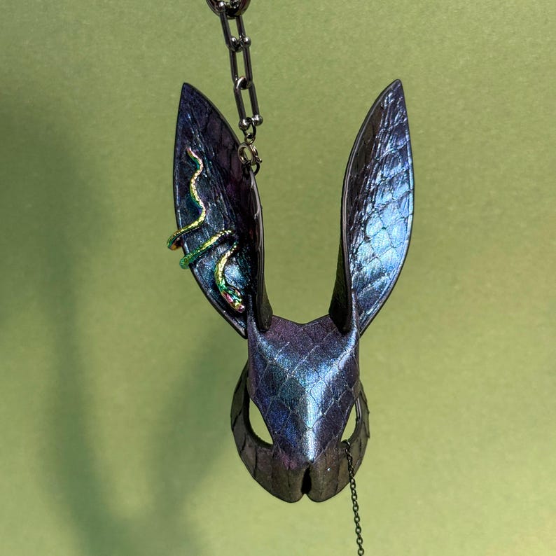 May include: An iridescent rabbit mask with a textured, scale-like pattern. The mask features long, upright ears and is decorated with a green and gold snake detail on one ear. A silver chain is attached to the mask.