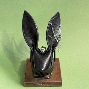 Op de afbeelding: Een zwart leren konijnenmasker met lange, rechtopstaande oren. Een kleine zilveren schedel siert het voorhoofd en een zilveren ketting bungelt aan &eacute;&eacute;n oor. Het masker is gemonteerd op een houten voet met een zwart metalen frame.