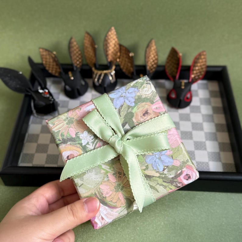 May include: A gift box with a floral pattern, tied with a pale green ribbon. Black rabbit figurines with gold and red details are displayed on a chequered tray. The background is a solid green.