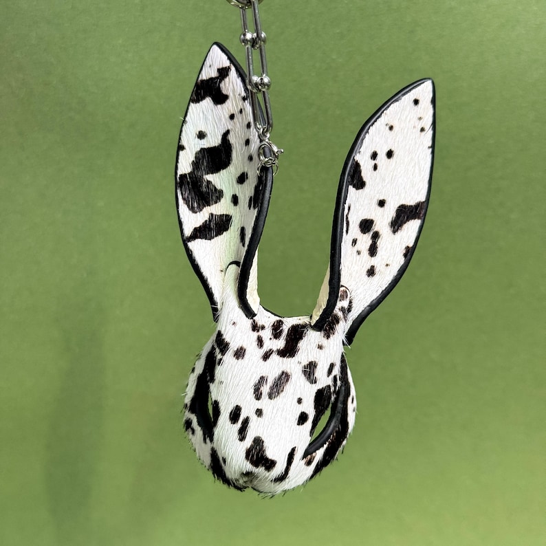 Pode incluir: Um chaveiro extravagante com uma cabe&ccedil;a de coelho e orelhas longas. O chaveiro &eacute; feito de material branco com manchas pretas, assemelhando-se a um padr&atilde;o d&aacute;lmata. As orelhas s&atilde;o contornadas em preto, e o chaveiro pende de uma corrente prateada.