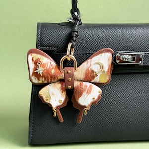 Pode incluir: Um pendente de mala em forma de borboleta com detalhes em castanho, branco e dourado. O pendente apresenta uma correia de couro e ferragens douradas. As asas têm um padrão de pele de vaca com detalhes dourados de estrela e lua. O pendente está preso a uma mala preta.