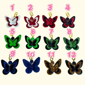 Twelve handmade leather wallet charm options displayed in a numbered grid, each in a different butterfly color design. All styles are crafted from layered genuine snakeskin and French lambskin with gold-plated hardware. The image shows color choices including red, white, black, blue, green, and brown patterns. These leather purse charms and butterfly zipper charms help shoppers select the exact style they want.