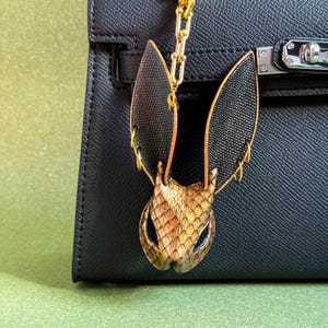 May include: A gold-toned metal and black leather bat-shaped bag charm with a gold chain. The charm is attached to a black leather handbag. The bat has textured wings and a detailed face.