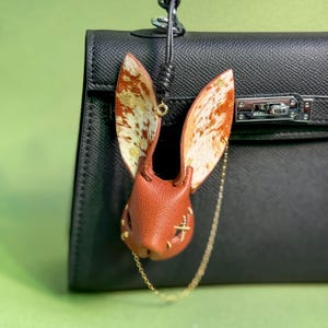 May include: A leather bunny charm with brown ears and a matching head, accented with gold-colored stitching and a gold chain. The charm is attached to a black handbag, creating a unique accessory.