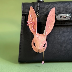 May include: A pink and gold rabbit mask charm with a dangling pink teardrop gem. The charm has a textured, scaly appearance and is attached to a black cord. A small pink tentacle-like detail is on the left ear.