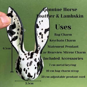 Pode incluir: Um pendente de orelhas de coelho em couro manchado a preto e branco. O pendente tem 6,5 cm de altura e 3,5 cm de largura. O texto diz "Genuine Horse Leather & Lambskin" e "Bag Charm, Keychain Charm, Statement Pendant, Car Rearview Mirror Charm."