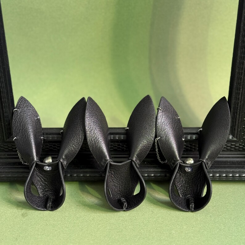 Op de afbeelding: Drie zwarte leren konijnenoren haarbanden worden gepresenteerd tegen een groene achtergrond. Elke haarband heeft een paar rechtopstaande oren met een klein kettingdetail. De haarbanden zijn gepositioneerd voor een zwart omlijst object.