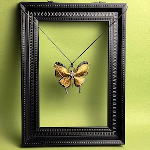 May include: A gold and black butterfly pendant hangs inside a black picture frame against a green background. The butterfly pendant has intricate details and is suspended by a thin black cord. The frame has a textured border.