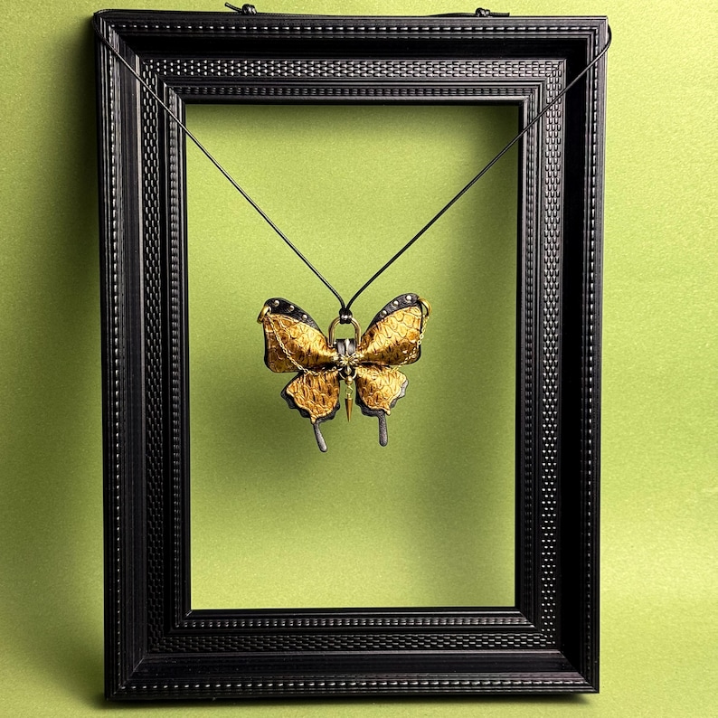 May include: A gold and black butterfly pendant hangs inside a black picture frame against a green background. The butterfly pendant has intricate details and is suspended by a thin black cord. The frame has a textured border.