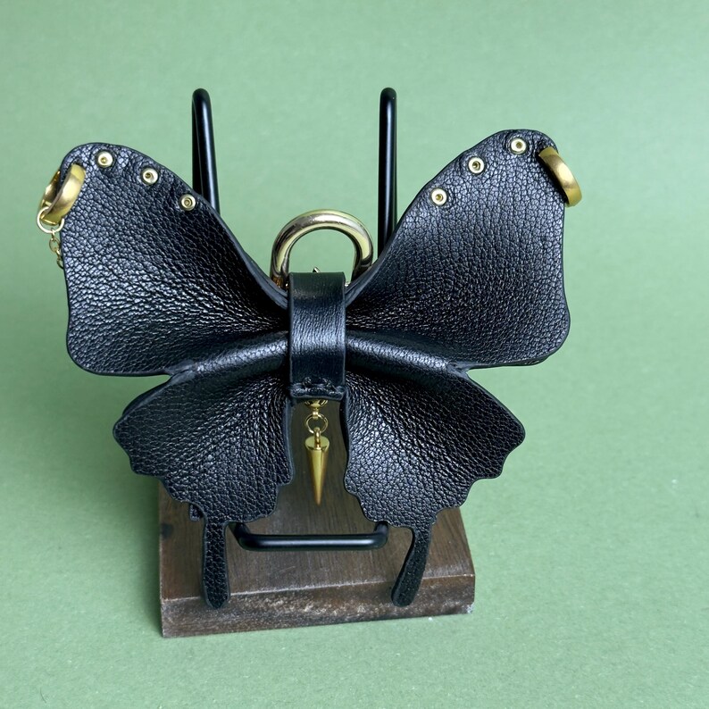 May include: A black leather butterfly-shaped keychain with gold-colored hardware. The butterfly has riveted details and a dangling gold-colored charm. The keychain is displayed on a wooden block with a black metal stand against a green background.