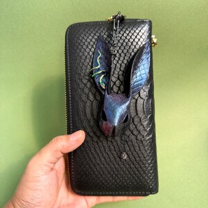 Black snakeskin-pattern long wallet held in one hand, featuring a handmade blue-purple rainbow leather bunny bag charm attached at the top. The bunny charm has tall textured snakeskin ears, iridescent multicolor highlights, and a small dangling crystal on a dark chain. The contrast between the dark wallet and the vibrant rainbow leather makes the charm stand out clearly.