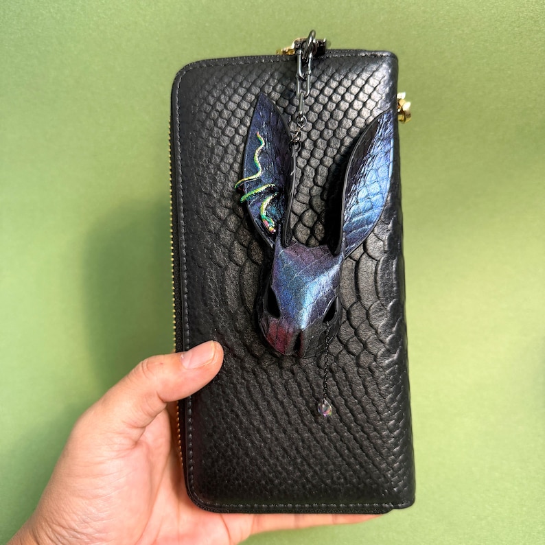 Black snakeskin-pattern long wallet held in one hand, featuring a handmade blue-purple rainbow leather bunny bag charm attached at the top. The bunny charm has tall textured snakeskin ears, iridescent multicolor highlights, and a small dangling crystal on a dark chain. The contrast between the dark wallet and the vibrant rainbow leather makes the charm stand out clearly.