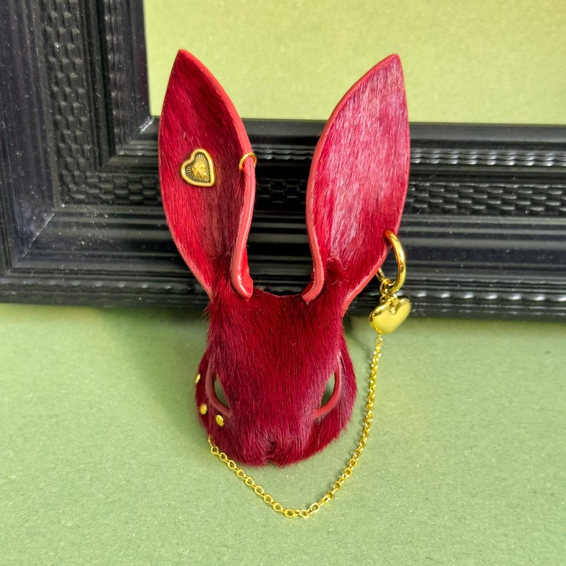 May include: A red rabbit head-shaped accessory with gold accents. The rabbit has long, upright ears and a textured fur-like appearance. Gold heart-shaped charms and a chain dangle from the ears. Small gold studs are placed around the eyes.