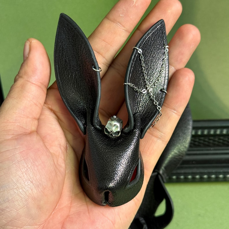 Handheld close-up of a goth luxury bunny leather bag charm made from handmade lambskin, showing detailed long ears, skull accent, and metal chain decoration. Dark gothic wicked purse charm displayed in hand to show size, texture, and craftsmanship, designed as a statement handbag charm for luxury bags and edgy styles.