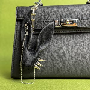Può includere: Una borsa in pelle nera con chiusura argentata e un ciondolo decorativo a forma di orecchie di coniglio nere. Il ciondolo ha punte argentate e una catena argentata. Lo sfondo è di colore verde.