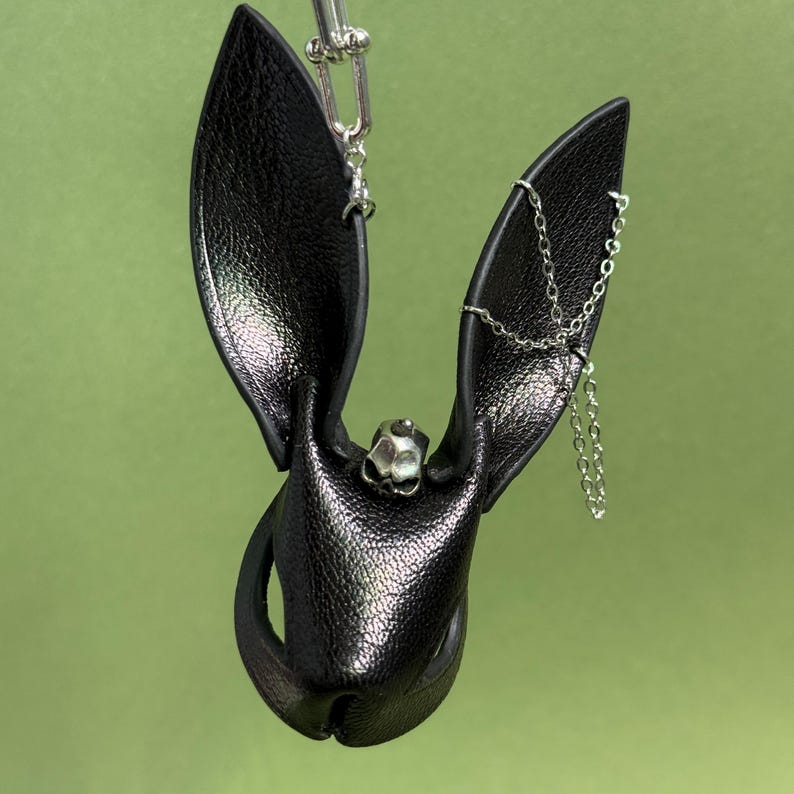 Op de afbeelding: Een zwart leren vleermuisvormig accessoire met een zilveren ketting en schedeldetail. De vleermuis heeft grote, puntige oren en een getextureerd oppervlak. Het accessoire is ontworpen om te worden gedragen of als decoratief item te worden gebruikt.