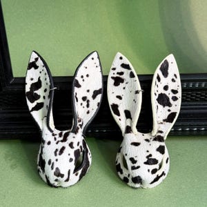 Pode incluir: Duas broches feitas &agrave; m&atilde;o em forma de orelhas de coelho. As broches s&atilde;o brancas com manchas pretas, assemelhando-se a um padr&atilde;o d&aacute;lmata. As orelhas est&atilde;o em p&eacute; e as broches t&ecirc;m uma apar&ecirc;ncia texturizada. Elas s&atilde;o colocadas sobre um fundo verde.