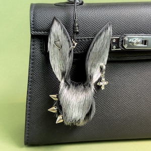 Peut inclure: Un porte-clés en forme d'oreilles de lapin noir et argenté, avec des pointes argentées, un charm en forme de cœur et un charm en forme de croix. Les oreilles de lapin sont en fausse fourrure et sont attachées à une lanière en cuir noir.