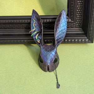 May include: A fantasy bunny mask with iridescent blue and purple scales. A small, colourful snake adorns one ear. A chain with a teardrop-shaped gem hangs from the mask's eye. The mask is set against a green background.