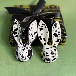 Pode incluir: Dois ornamentos de cabe&ccedil;a de coelho manchados em branco e preto com orelhas compridas. As cabe&ccedil;as de coelho est&atilde;o em frente a um presente embrulhado com um la&ccedil;o preto. O papel de embrulho &eacute; preto com detalhes dourados. O fundo &eacute; verde liso.