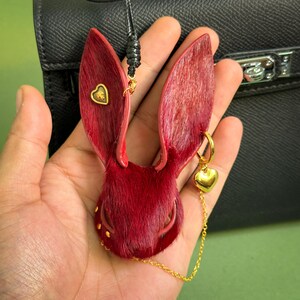 Close-up of a handmade Burgundy Gothic bunny leather bag charm shown in hand, featuring genuine horsehair texture, French lambskin backing, gold heart accents, and a delicate chain. Luxury Dark Romance purse charm designed for handbags and designer accessories.