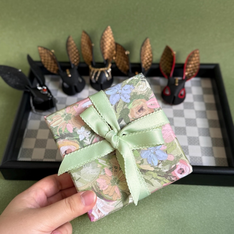 May include: A floral-patterned gift box tied with a light green ribbon. Several black rabbit figurines with gold and red accents are arranged on a checkered tray. The background is a solid green.