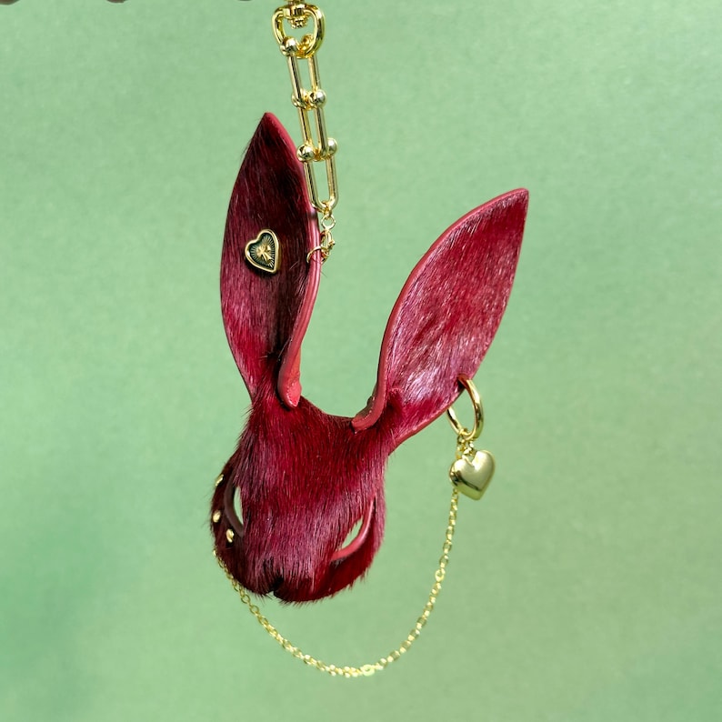 May include: A burgundy rabbit ear-shaped pendant with a gold chain and accents. The pendant has a gold heart-shaped charm and a small heart-shaped detail on one ear. The background is a solid green.