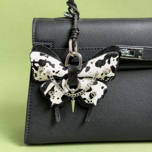 May include: A black and white butterfly-shaped bag charm with silver-tone hardware. The charm features a cowhide pattern on the wings, studded accents, and a dangling chain. It is attached to a black leather handbag.