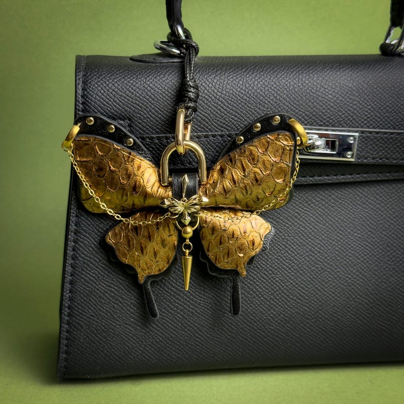 Gold vintage gothic butterfly leather bag charm handmade from luxury snakeskin and lambskin leather, featuring gold-tone metal hardware, chain accents, and a layered butterfly design. This leather butterfly purse charm is shown attached to a structured black handbag, designed as a statement handbag charm with a dark luxury vintage gothic aesthetic.