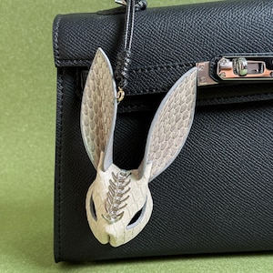 Puede incluir: Un charm de bolso decorativo con forma de conejo, con cara y orejas texturizadas de color blanco roto, detallado con un acento plateado. El charm cuelga de una correa de cuero negro, sujeta a un bolso de mano de cuero negro.